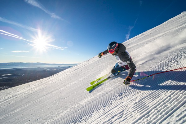 A skier carving a sharp turn down a bright, sunny slope with fresh powder, blue sky, and a blazing sun over the horizon.